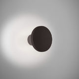 Arid A/01 round LED wall light in Textured Black aluminium, fluted disc on a short stem casting a soft halo of warm light onto a plain grey wall.