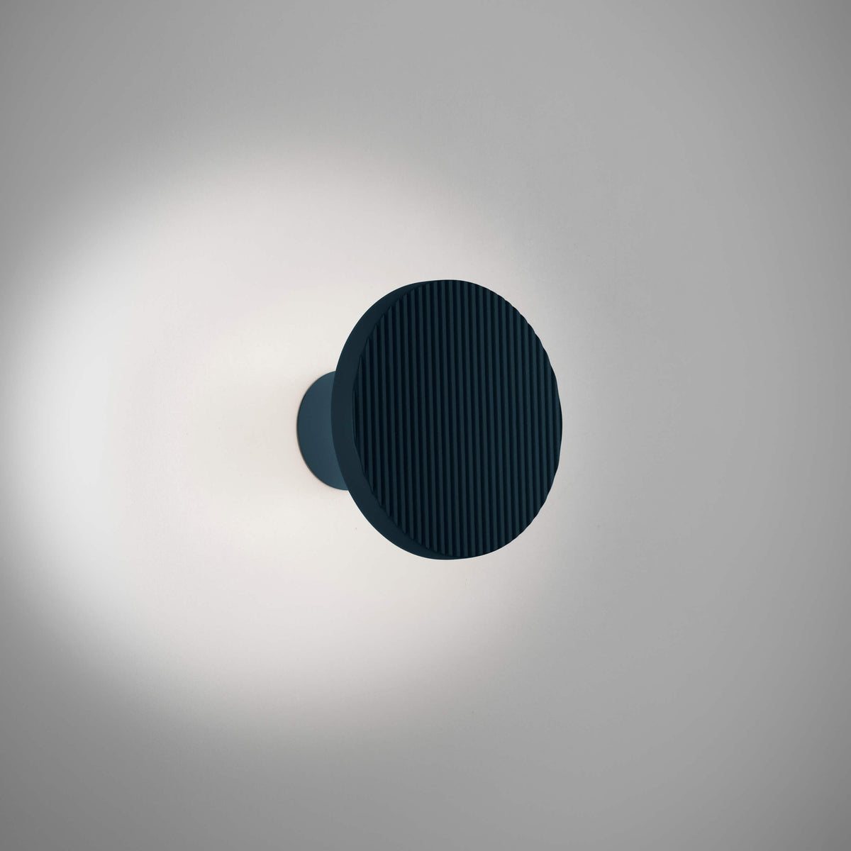 Arid A/01 round aluminium LED wall light with fluted disc face in Textured Grey Blue, mounted on a short stem, glowing softly against a plain light grey background.