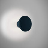 Arid A/01 round aluminium LED wall light with fluted disc face in Textured Grey Blue, mounted on a short stem, glowing softly against a plain light grey background.