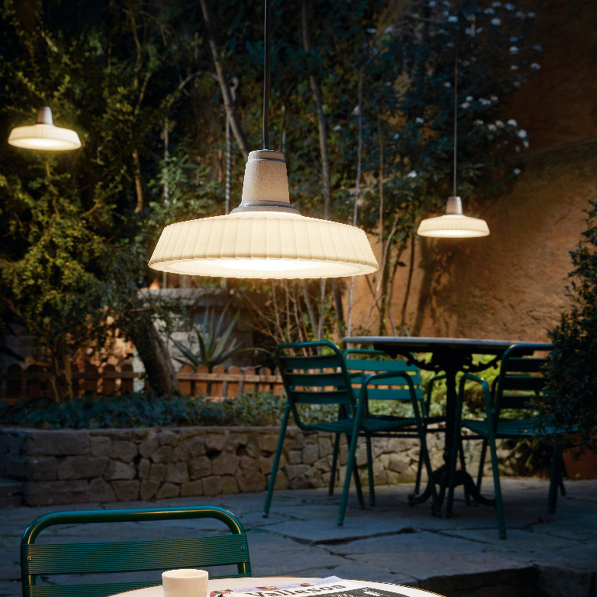 Marietta S/32 Dome LED Outdoor Pendant Light in white, with a pleated dome shade on a black cable, shown illuminated above a garden dining table at night.
