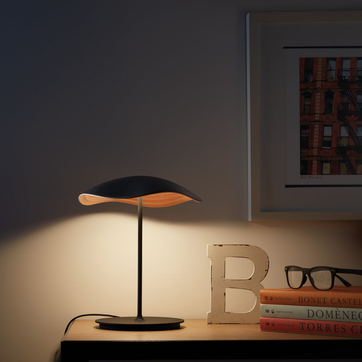 Valentina M/29 Table Lamp with wavy black and copper shade on a slim black stem and round base, glowing warmly on a wooden shelf beside books and glasses.