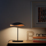 Valentina M/29 Table Lamp with wavy black and copper shade on a slim black stem and round base, glowing warmly on a wooden shelf beside books and glasses.