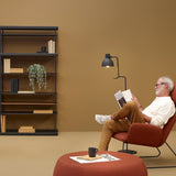 Blux F50 Aluminium Dome Floor Lamp in black with round base and jointed arm, angled beside a man reading in a rust armchair in a living room.
