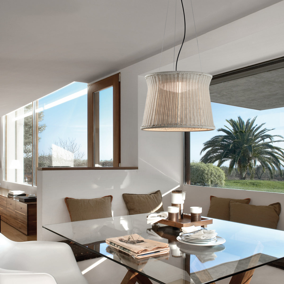 Syra 45 Aluminium LED Pendant Light with a woven ivory-white drum shade narrowing at the waist, hung above a glass dining table in a bright room with palm tree views.