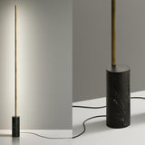 Hilow Line Nero Marquina linear floor lamp with slim brushed brass rod and black, white-veined marble cylindrical base, shown lit against a plain grey background and in close-up detail.