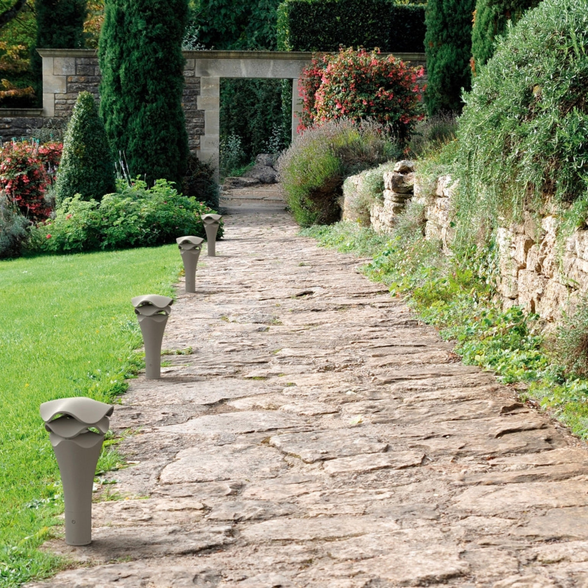 Cornet B/52 Cone LED Bollard Light in stone grey lining a stone garden path, showing its slender flared cone form with a wavy layered cap.