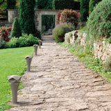 Cornet B/52 Cone LED Bollard Light in stone grey lining a stone garden path, showing its slender flared cone form with a wavy layered cap.