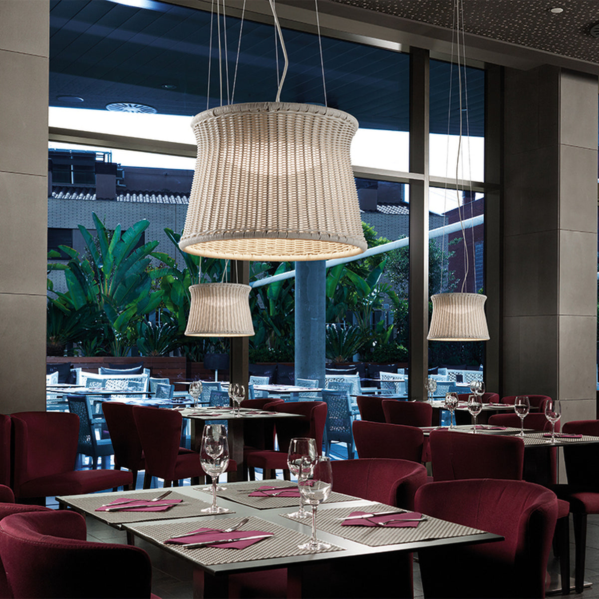 Syra 60 Aluminium LED Outdoor Pendant Light with woven ivory drum shade, shown above burgundy chairs in a restaurant dining area near large windows.