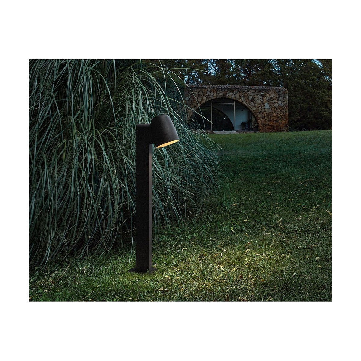 Nut B/60 Bell LED bollard light in textured black, with a bell-shaped head angled downward on a slim square post, glowing warmly in a garden lawn setting at dusk.
