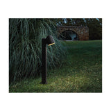 Nut B/60 Bell LED bollard light in textured black, with a bell-shaped head angled downward on a slim square post, glowing warmly in a garden lawn setting at dusk.