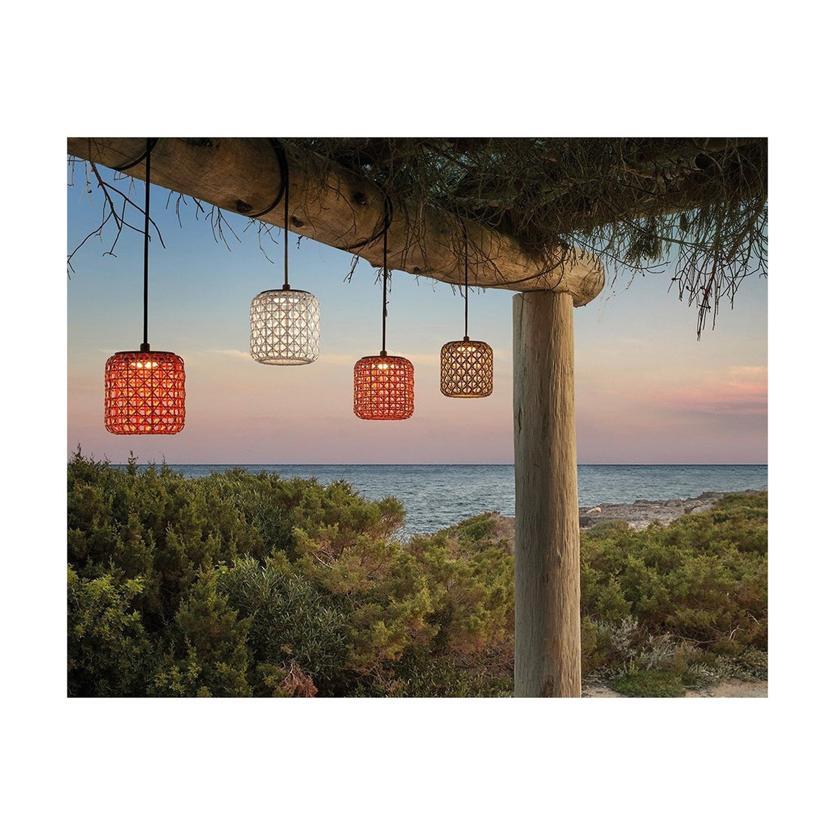 Nans S/16 woven cylindrical outdoor pendant light in brown, shown lit alongside white and terracotta shades hanging from a wooden pergola beam at dusk by the sea.