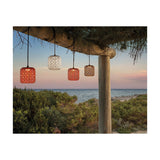 Nans S/16 woven cylindrical outdoor pendant light in brown, shown lit alongside white and terracotta shades hanging from a wooden pergola beam at dusk by the sea.