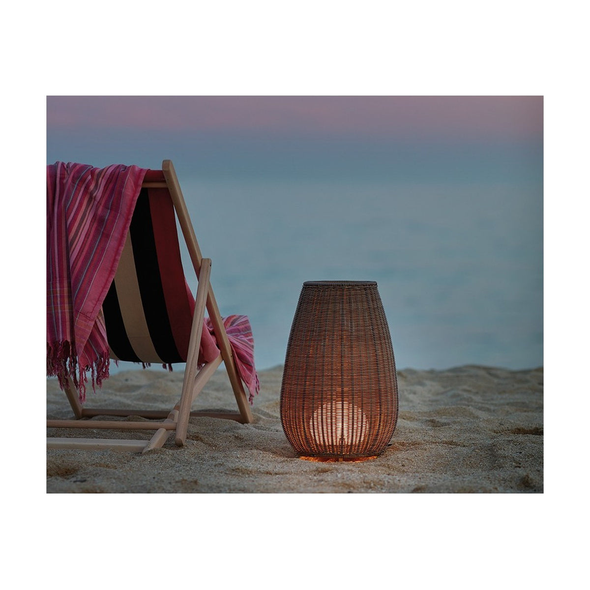 Amphora Woven Linear LED Outdoor Floor Lamp in beige, tapering woven shade glowing warm white, set on sandy beach beside a wooden deck chair at dusk.