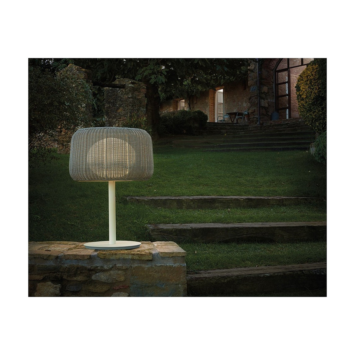 Fora M Drum LED Outdoor Table Lamp in Light Beige with woven drum shade on a slim white stem and round base, glowing on a garden wall beside stone steps and lawn at dusk.