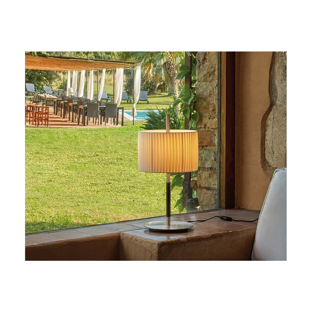 Danona T Ribbon Drum Table Lamp with pleated white drum shade on a slim satin nickel stem and round base, shown on a windowsill beside a garden and pool view.