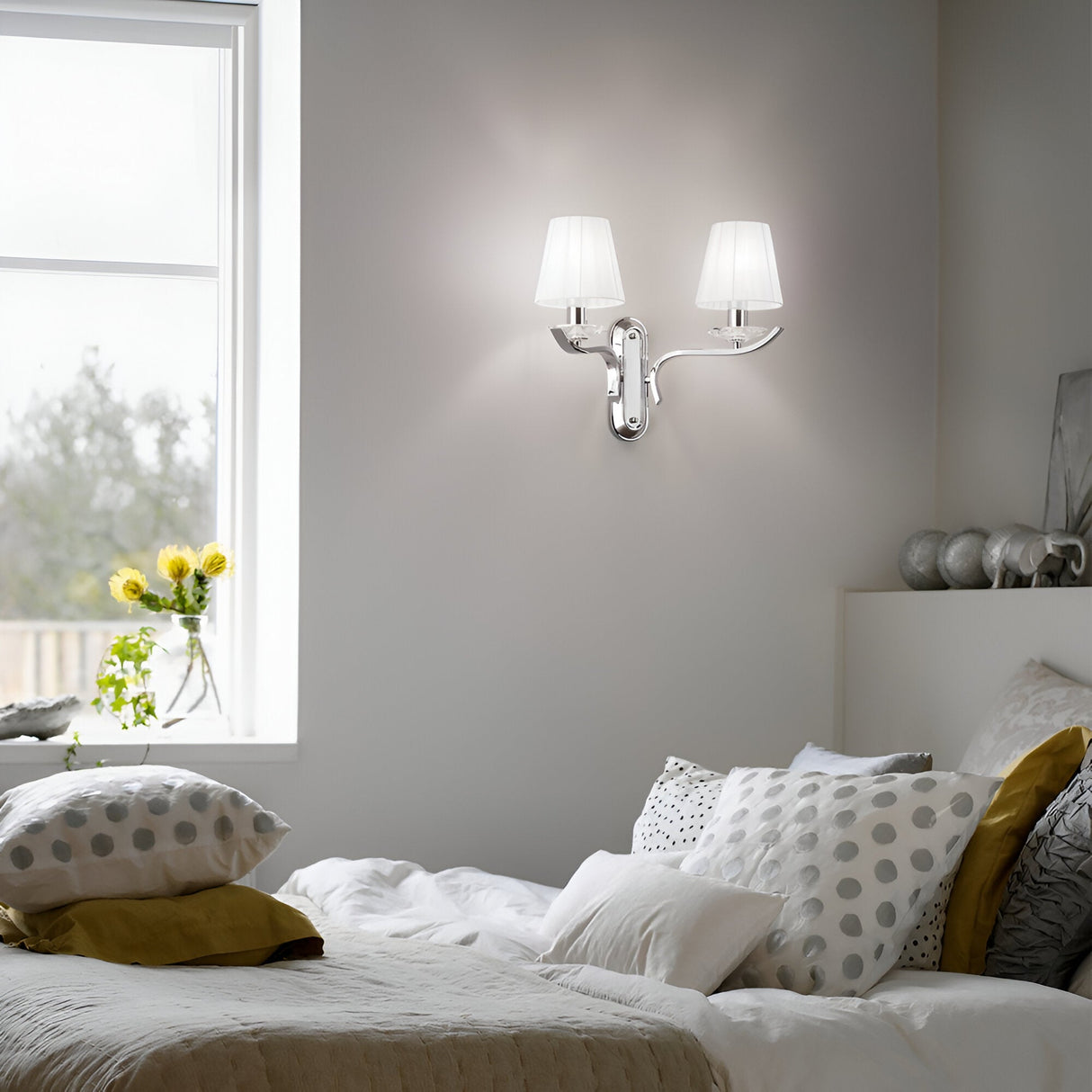 In the cozy bedroom, a white bed is accentuated with yellow and gray pillows under a Solum 2 Light Wall Light - White, showcasing its chromed metal frame and organza lampshade. The window reveals a view of vibrant greenery, enhanced by a small vase of yellow flowers on the sill.