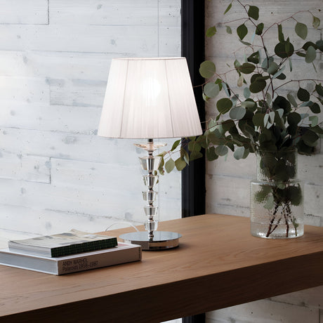 The Solum Large Crystal Table Lamp with its white organza lampshade graces a wooden table beside a vase filled with eucalyptus branches. A stack of art books complements the tranquil backdrop of light-colored textured walls.