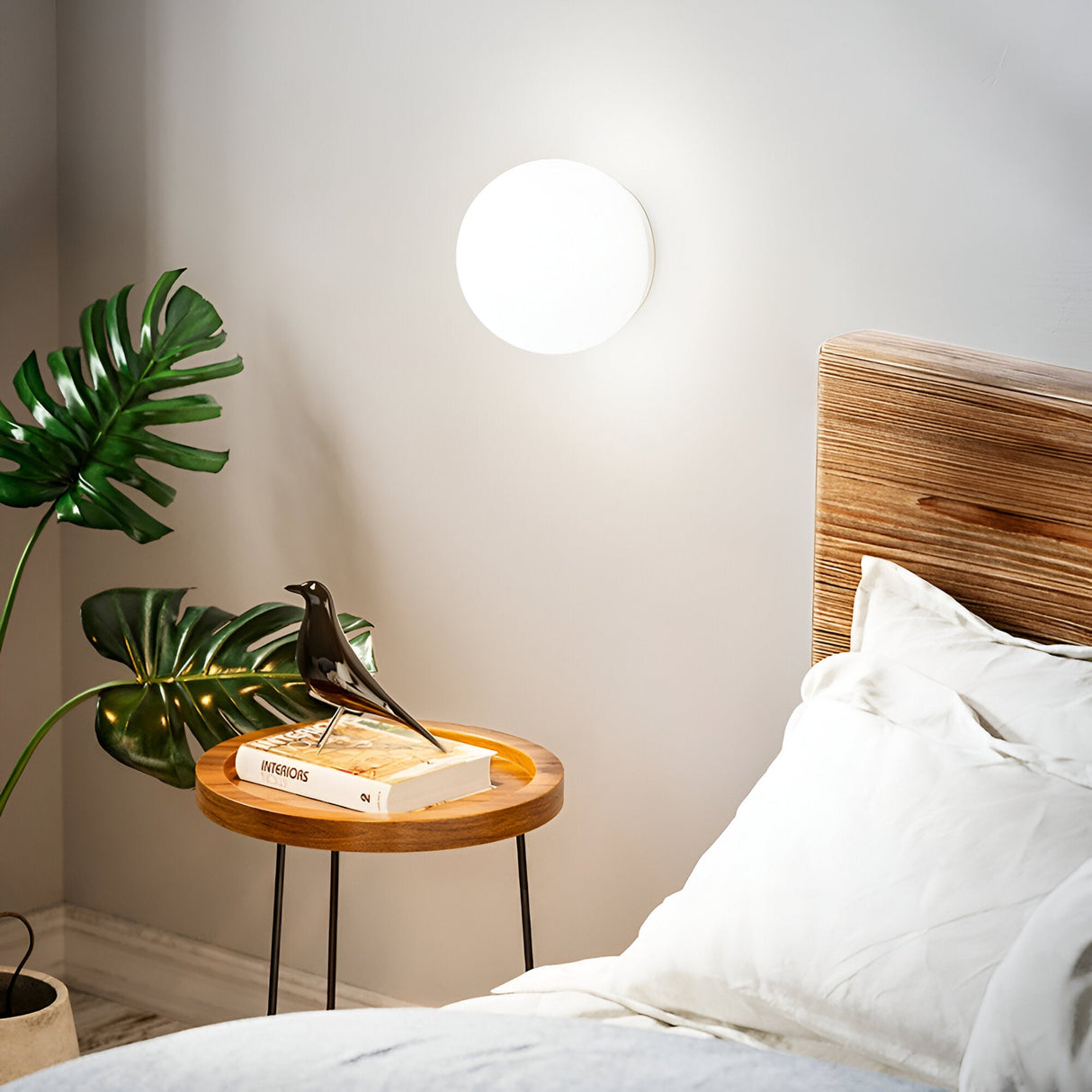 A modern bedroom scene features a wooden side table beside the bed. It showcases books, a small black decorative bird, and is softly illuminated by a Lyria Wall Light - White, 20cm, enhancing the room's ambient lighting. A green leafy plant sits alongside, completing the modern design.