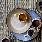 Sandrine Stoneware Deep Plate in light grey with speckled glaze and dark rim, shown holding a mustard mug on a woven tabletop setting.