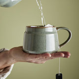 Hand holding a green stoneware Pixie cup with an open loop handle while hot water is poured in over a tea bag, set against a plain olive background.