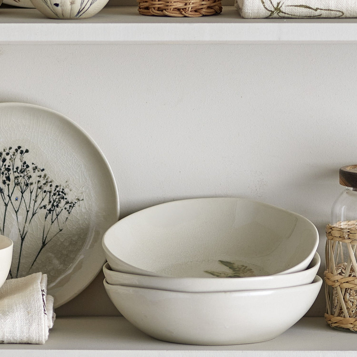 Bea Crackle Stoneware Deep Plates in cream, stacked on a shelf with an irregular rim, crackled finish, and a faint fern leaf motif, beside a matching patterned plate.