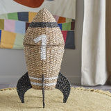 Cosmina Rocket Seagrass Basket with Lid in natural woven finish with black painted bands and a white "1", displayed on a rug in a child's room.