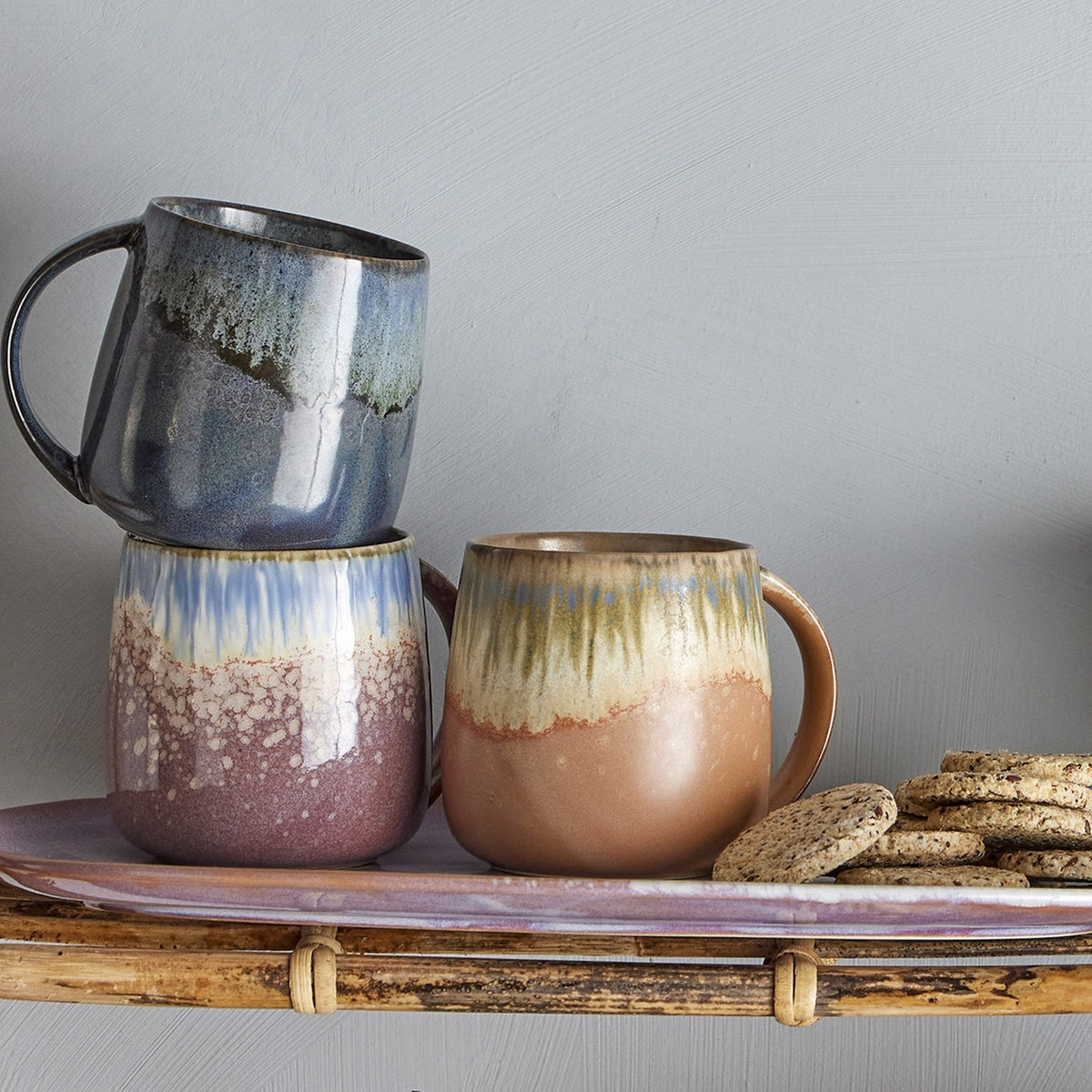Cloe Speckled Stoneware Mugs, set of 3, in blue, pink and brown reactive glaze with speckled lower bodies, shown on a tray with biscuits against a grey wall.