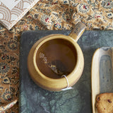 Solange Stoneware Mug 350ml in green with tea and loop handle, shown on a marble tray beside patterned fabric and a biscuit.
