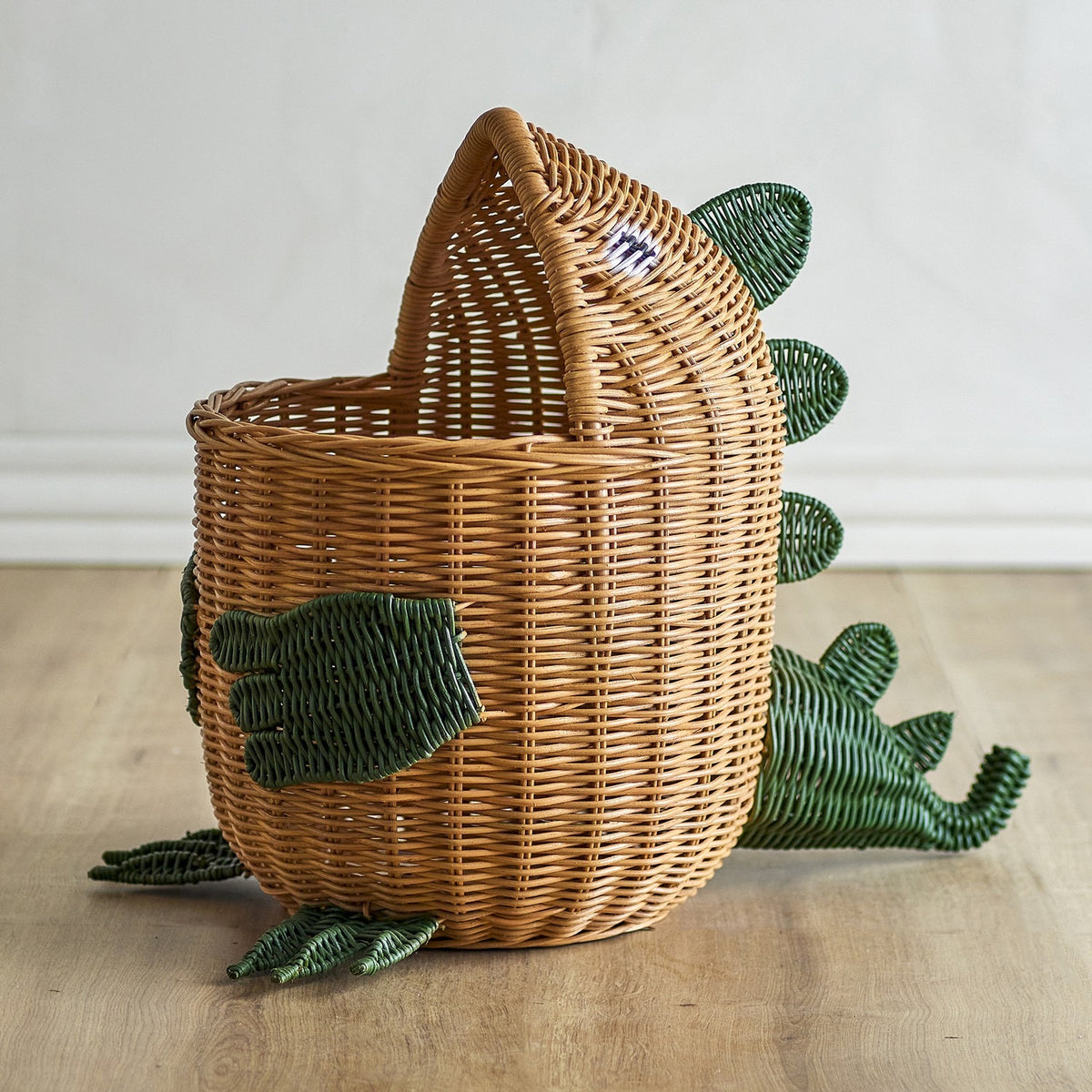 Eddi Dinosaur Rattan Basket in natural woven rattan, shaped with a rounded body, raised head, curved tail and green spines, arms and clawed feet, set on a wooden floor.