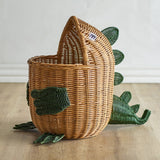 Eddi Dinosaur Rattan Basket in natural woven rattan, shaped with a rounded body, raised head, curved tail and green spines, arms and clawed feet, set on a wooden floor.