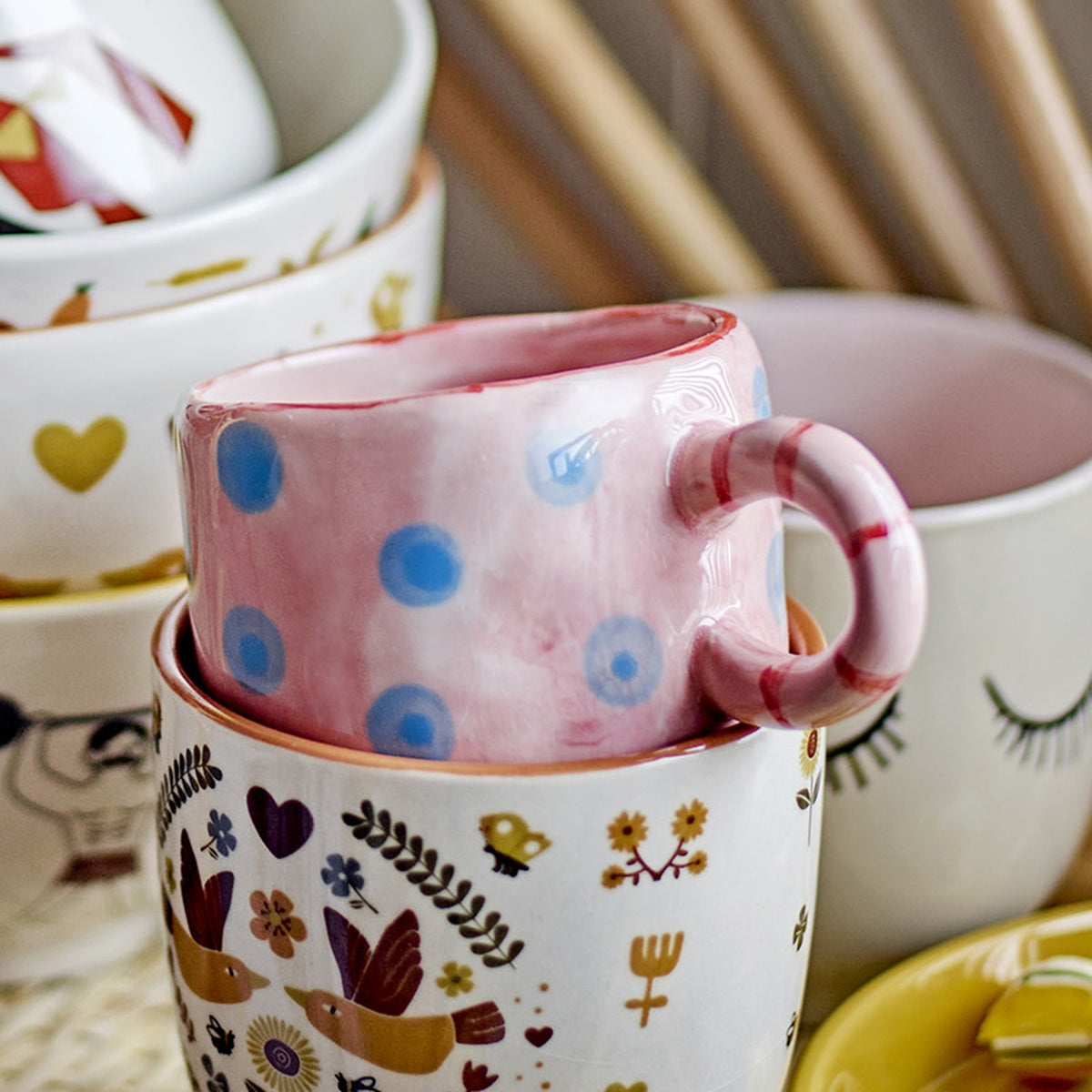 Nini Dotted Stoneware Cup in pink with blue polka dots and a striped ring handle, nestled inside a patterned ceramic mug among other decorative cups.
