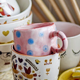Nini Dotted Stoneware Cup in pink with blue polka dots and a striped ring handle, nestled inside a patterned ceramic mug among other decorative cups.