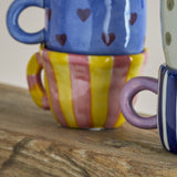 Nini Yellow Striped Stoneware Cup 110ml in multicoloured pink and yellow stripes with a loop handle, stacked with other patterned mugs on a wooden surface.