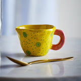 Nini Stoneware Cup 100ml in yellow with hand-painted green spotted pattern and a coral pink loop handle, shown with a gold spoon on a marble surface.