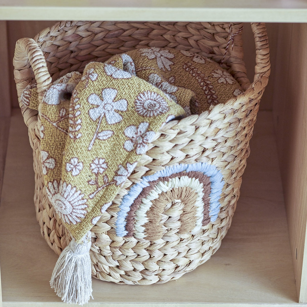 Agnes Water Hyacinth Basket in beige with woven handles and a stitched rainbow motif, holding a folded mustard floral cloth on a shelf.