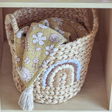 Agnes Water Hyacinth Basket in beige with woven handles and a stitched rainbow motif, holding a folded mustard floral cloth on a shelf.