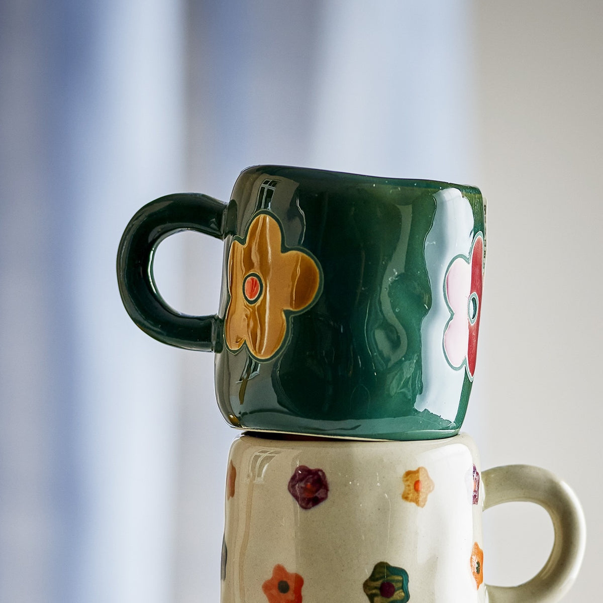 Addy Stoneware Cup 130ml in green, a small ceramic kids' cup with loop handle and bold flower motifs, stacked atop a floral-patterned mug against a blurred light background.