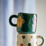 Addy Stoneware Cup 130ml in green, a small ceramic kids' cup with loop handle and bold flower motifs, stacked atop a floral-patterned mug against a blurred light background.