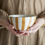 Hands holding the Begonia Striped Stoneware Bowl 14cm in Brown, a white ceramic bowl with hand-painted brown vertical stripes running from base to rim.