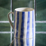 Begonia Striped Stoneware Mug 440ml in Blue, handle to the left, hand-painted blue vertical stripes on white ceramic, set against a green tiled background.