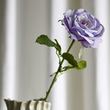 Artificial rose stem with a single open purple bloom and ruffled petals, serrated green leaves along the stem, displayed in a white ridged vase against a soft curtain backdrop.