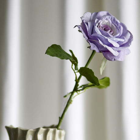 Artificial rose stem with a single open purple bloom and ruffled petals, serrated green leaves along the stem, displayed in a white ridged vase against a soft curtain backdrop.
