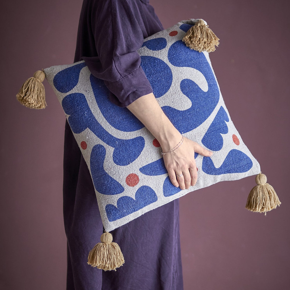 Eviaya Cotton Cushion in Multi, square with bold blue abstract shapes and red dots on cream, tasselled corners, held under a woman's arm against a plum background.