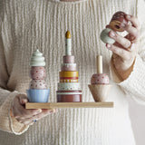 Celebrate 18-Piece Dotted Wooden Activity Toy held by hand over a wooden base, showing stacked dessert towers with painted sprinkles in pink, blue, green and brown tones.
