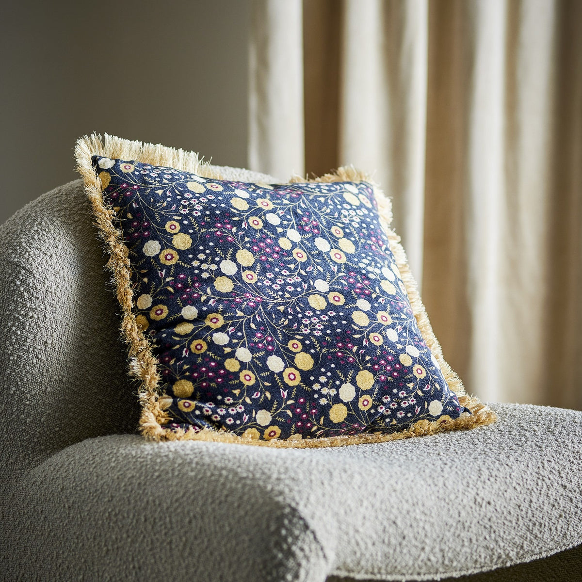 Eydis Floral Cushion in Multi, a square fringed cushion with a dense navy floral print in gold, pink and white, shown on a grey upholstered armchair.