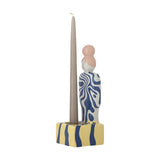 Priya Stoneware Tealight Holder in Multi, a ceramic figure with a blue swirl-patterned body and pink bun, holding a tall taper candle on a yellow striped base, shown on a white background.