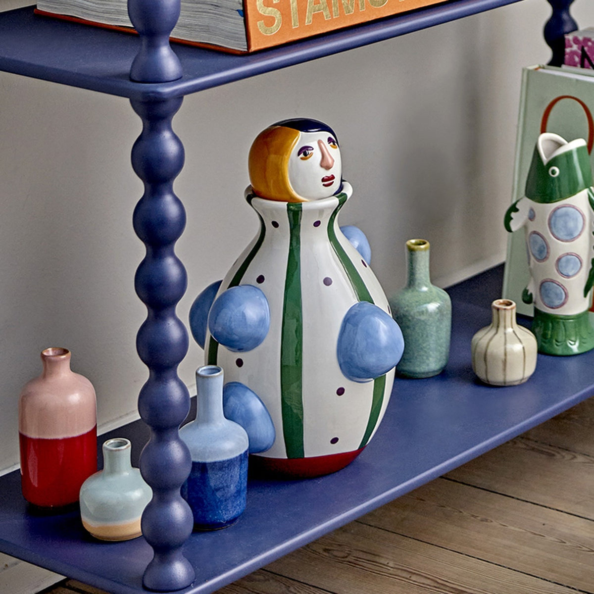 Conneli Ceramic Vase 33cm in Multi, a figure-shaped vase with painted face, striped and dotted body, and raised blue knobs, shown on a navy shelf among smaller vases.