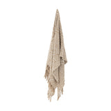 Okapi Cotton Throw in beige, hanging with a woven wavy-line pattern and fringed edges, against a plain white background.