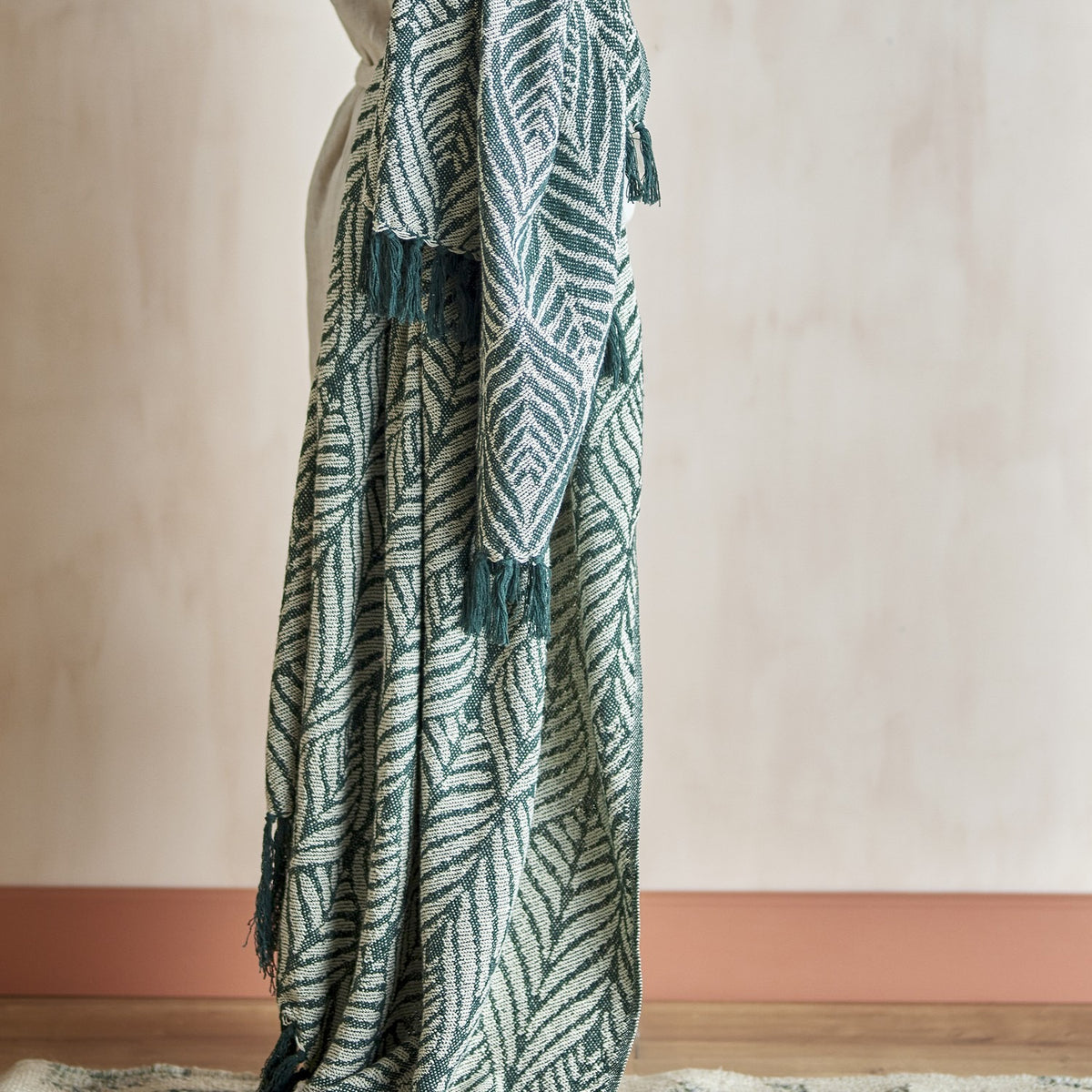 Ugento Leaf Cotton Throw in green, woven with a bold leaf pattern and knotted fringe, draped over a person's arm against a plain neutral wall.