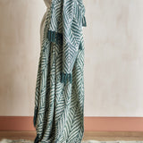 Ugento Leaf Cotton Throw in green, woven with a bold leaf pattern and knotted fringe, draped over a person's arm against a plain neutral wall.
